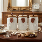 3 piece white ceramic canister set with geometric embossed design, bamboo lids, leather loop handles, labeled tags, and a wooden tray for kitchen storage.
